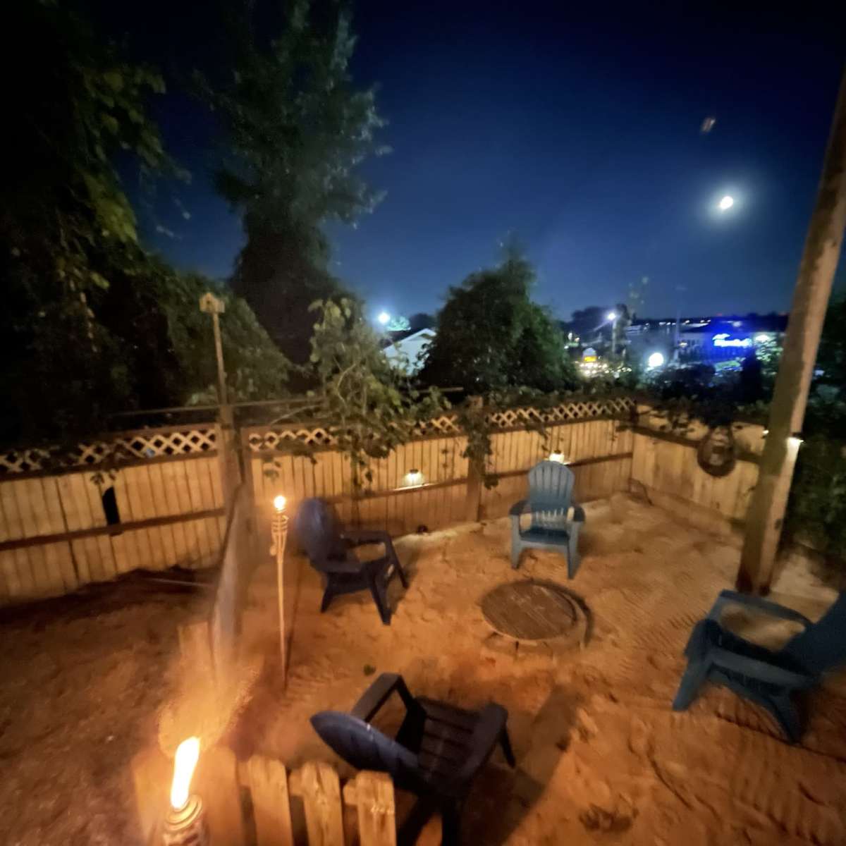 A nighttime outdoor seating area with four blue chairs arranged around a fire pit, surrounded by trees and a fence.