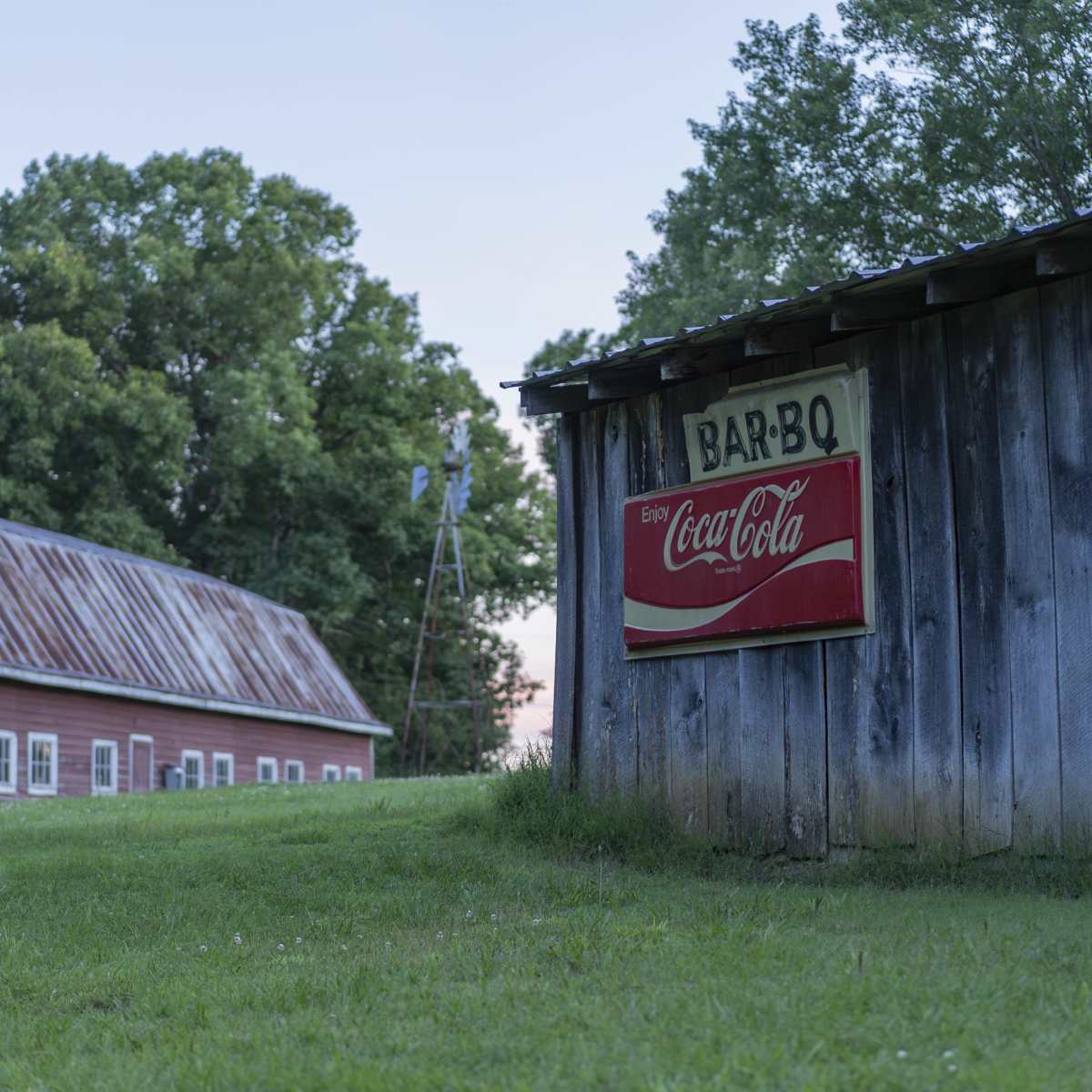 A weathered barn with a Coca-Cola sign stands next to a red building in a grassy field.
