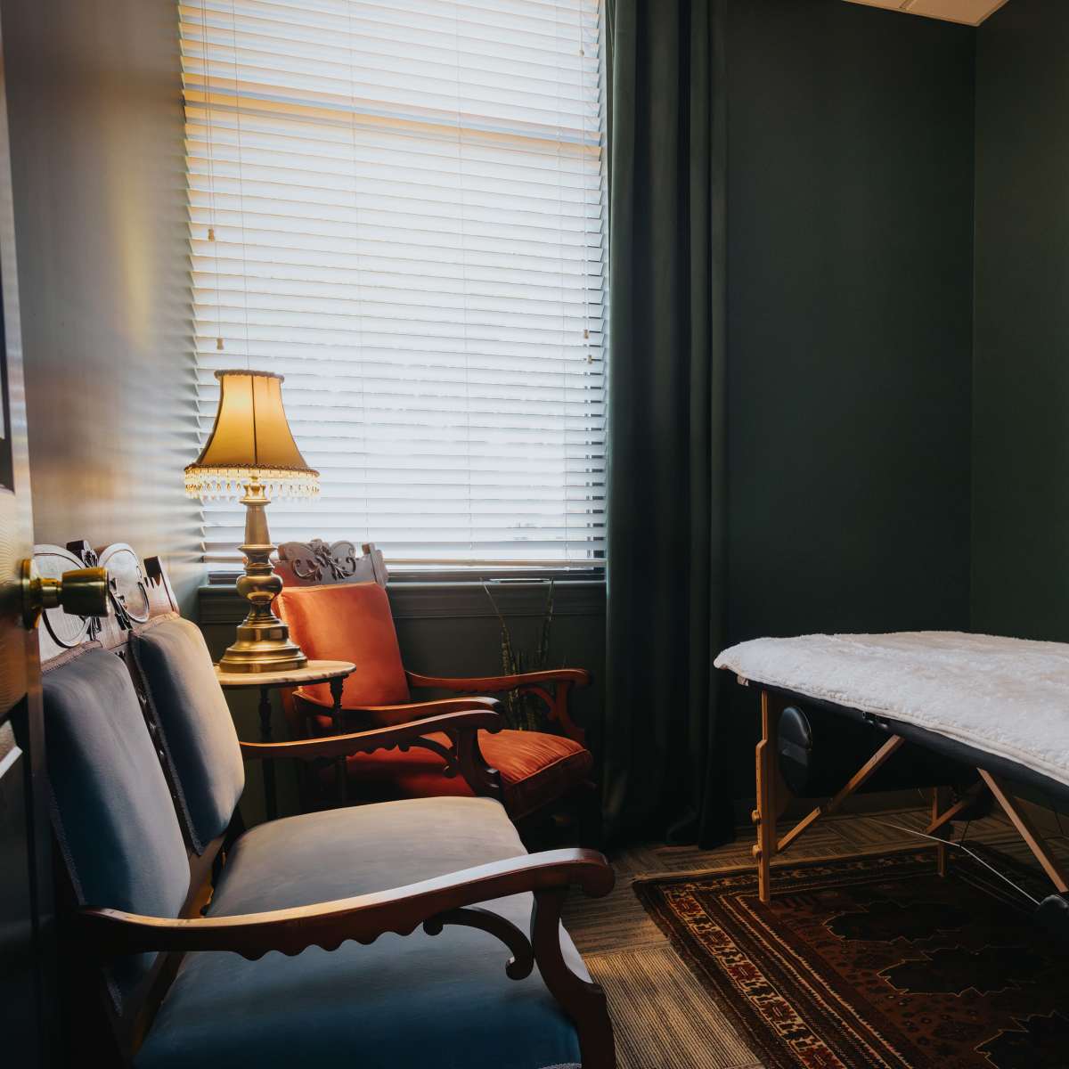 A massage room features a massage table covered with a white blanket, two upholstered chairs, a lamp, and artwork on the walls.