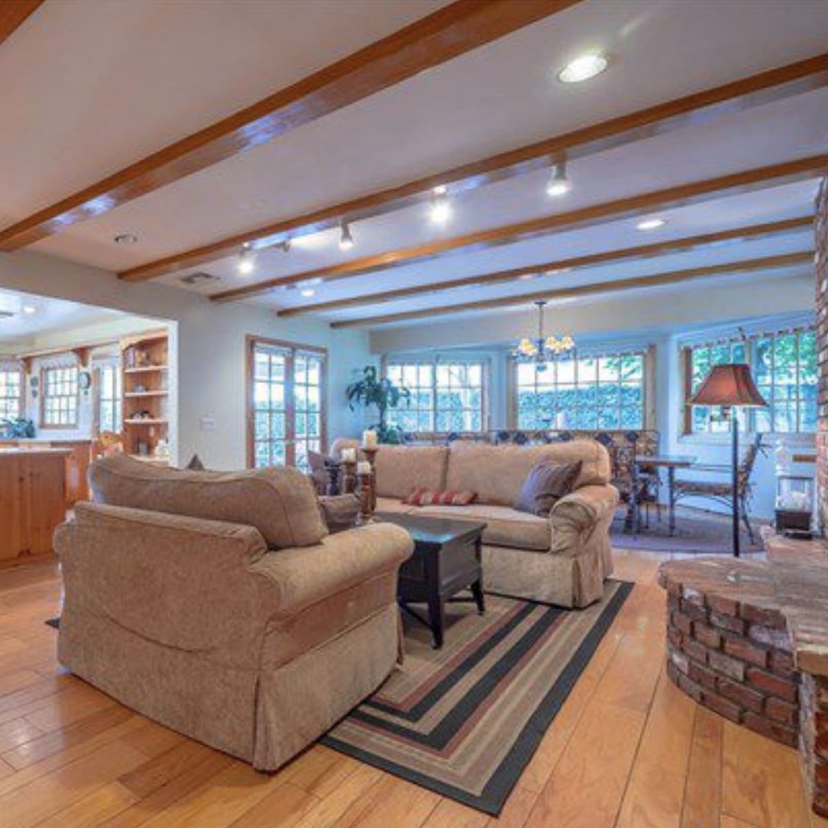 A living room features two sofas facing a black coffee table, with wooden beams on the ceiling and a view into a kitchen area with light wooden cabinetry.