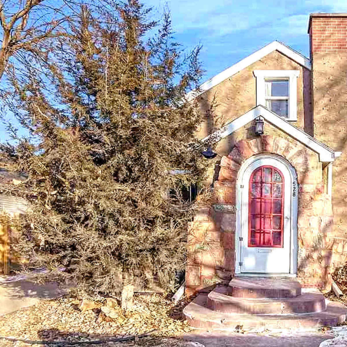 A single-story house with a stone archway around the front door and a tall evergreen tree nearby.