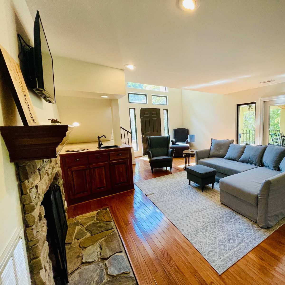 A living room with a large gray sectional sofa, a stone fireplace, and a bar area, featuring large windows that provide a view of the outdoors.