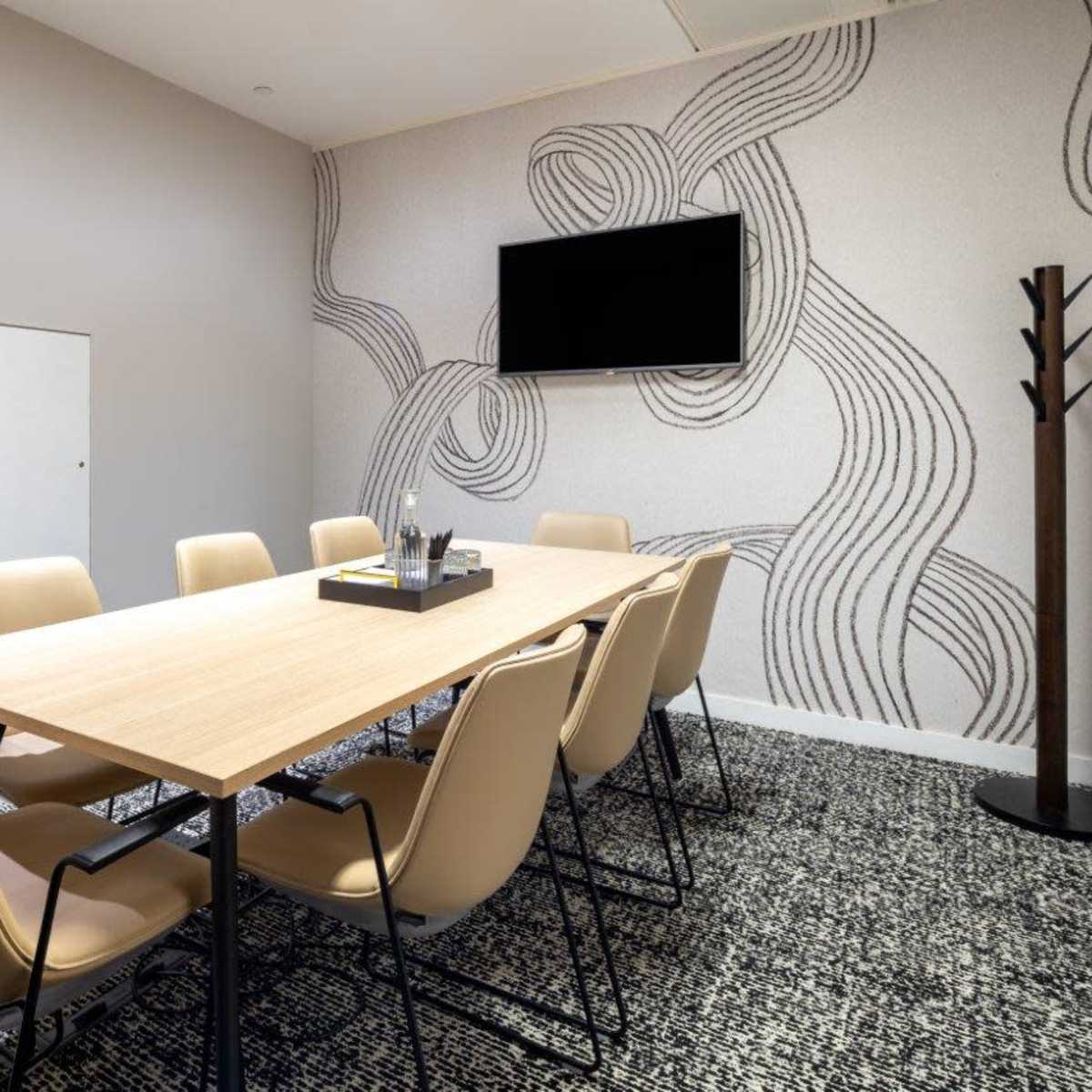A modern conference room features a long wooden table surrounded by beige chairs, with a wall-mounted television and a decorative patterned wall.