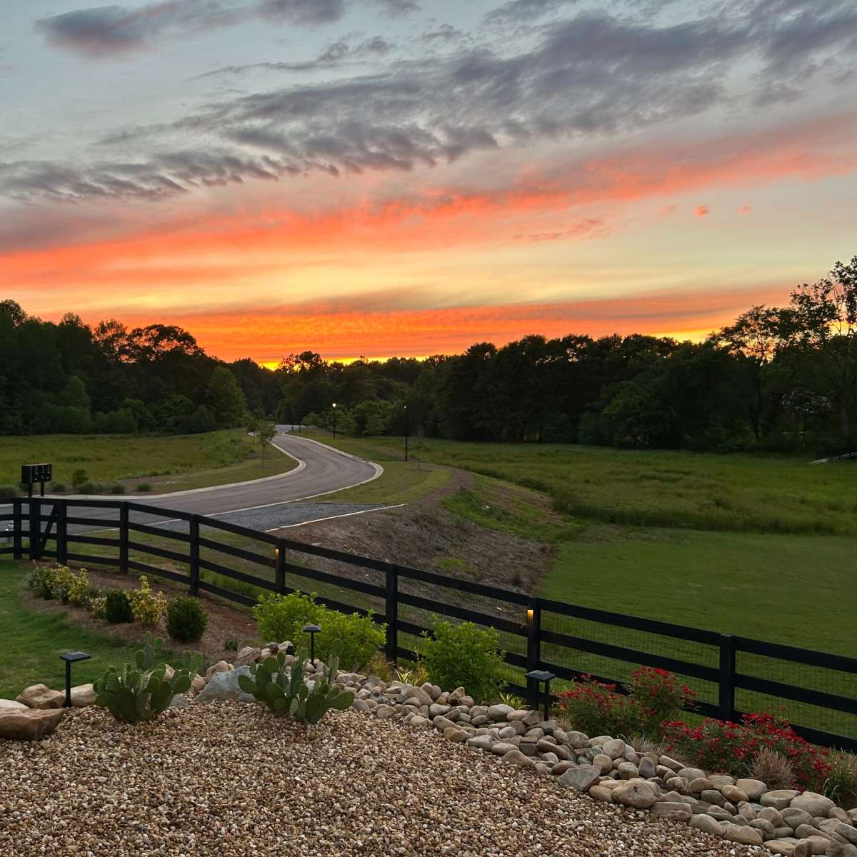 The scene shows a winding road bordered by a black fence, with a vibrant sunset casting colors across the sky and a landscaped foreground featuring rocks and plants.