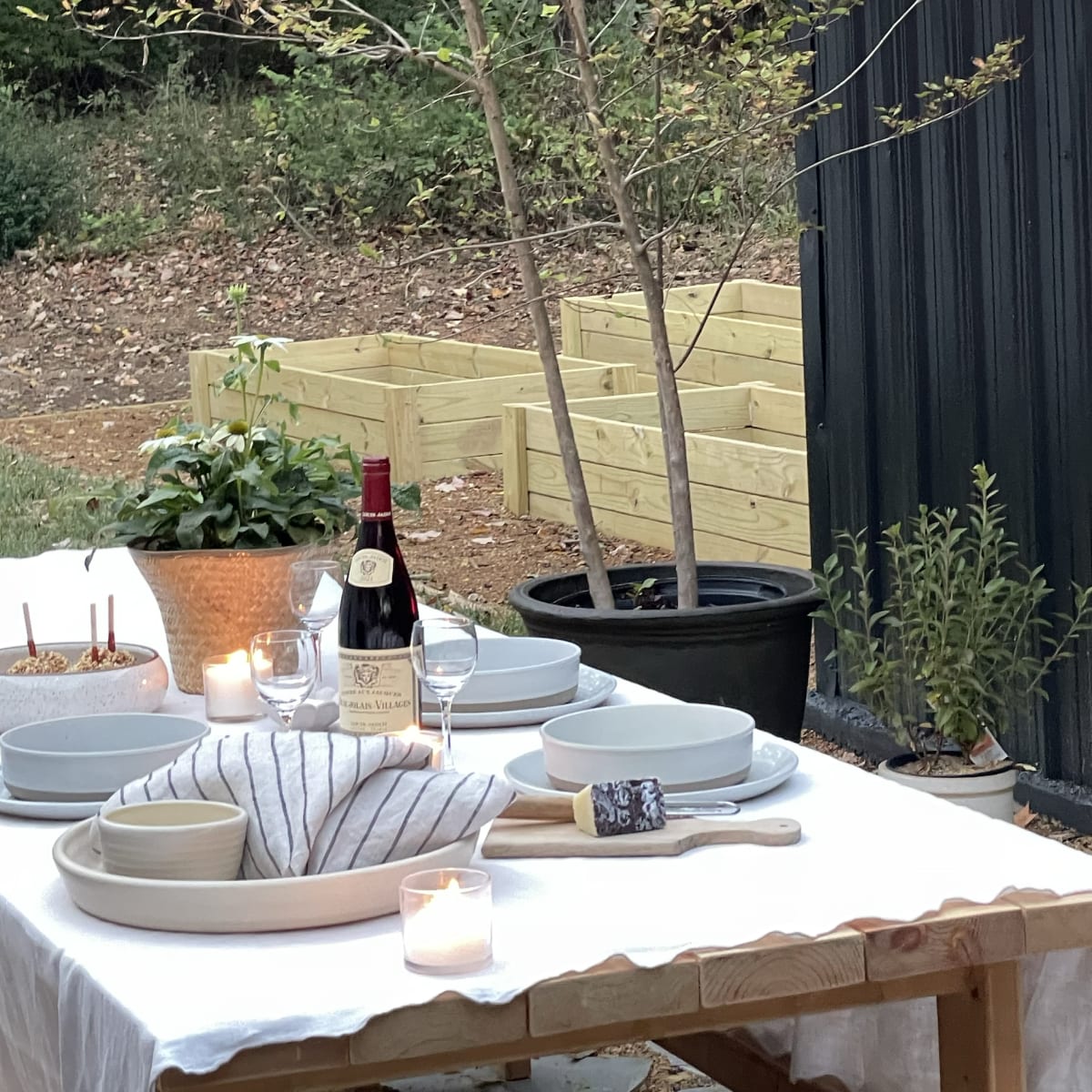 An outdoor dining table is set with plates, glasses, and a bottle of wine, beside potted plants and a garden bed.