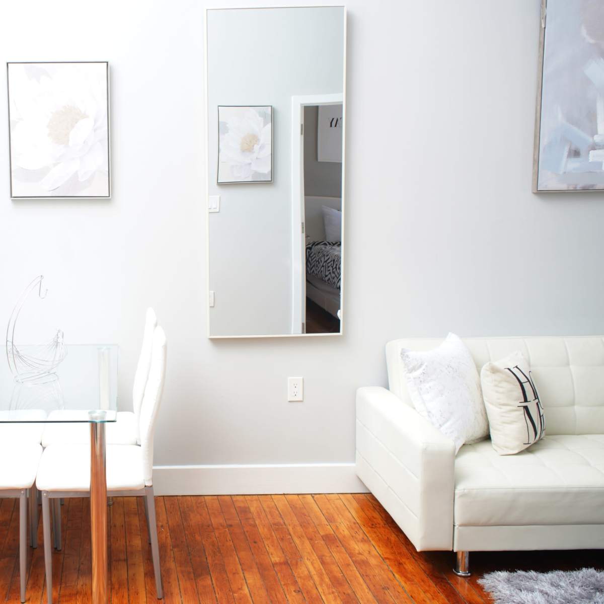 A glass dining table with white chairs is positioned beside a large mirror and framed artwork in a light-colored room with wooden flooring.