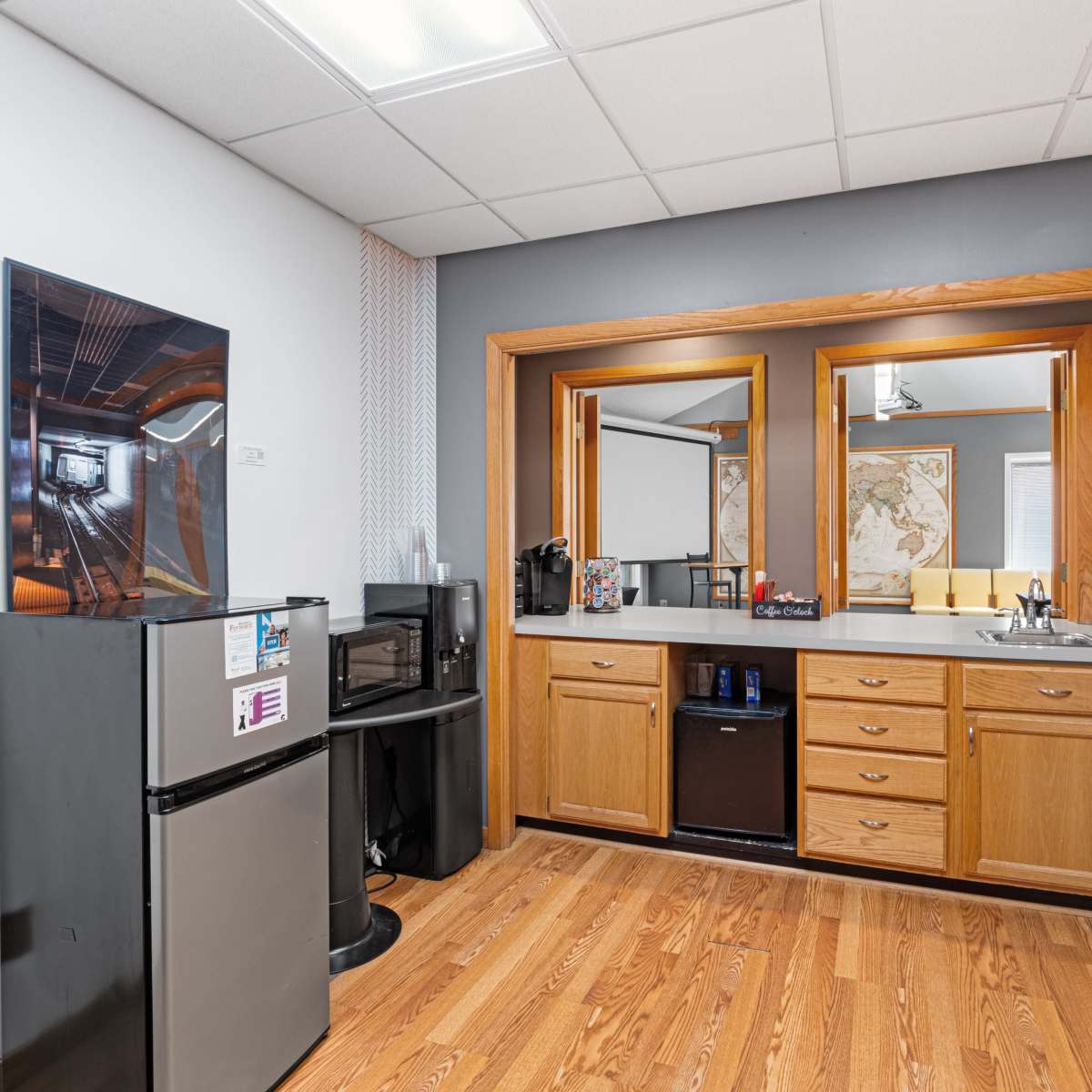 A kitchenette area with wooden cabinets, a small refrigerator, a microwave, and a countertop, separated from a room with a map on the wall.