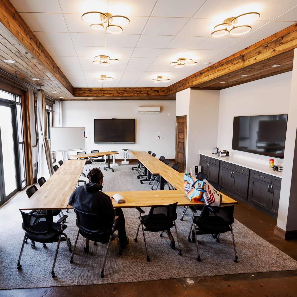 A modern conference room features a long wooden table surrounded by black chairs, with large windows and two screens mounted on the walls.