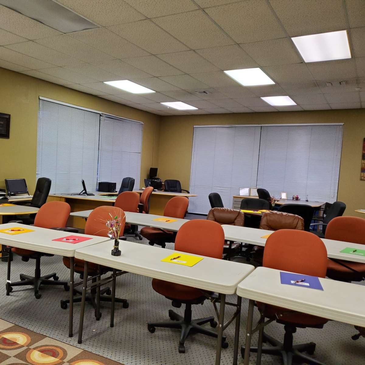 A classroom or training room setup with several tables and chairs, computers on desks, and colorful papers placed on the tables.