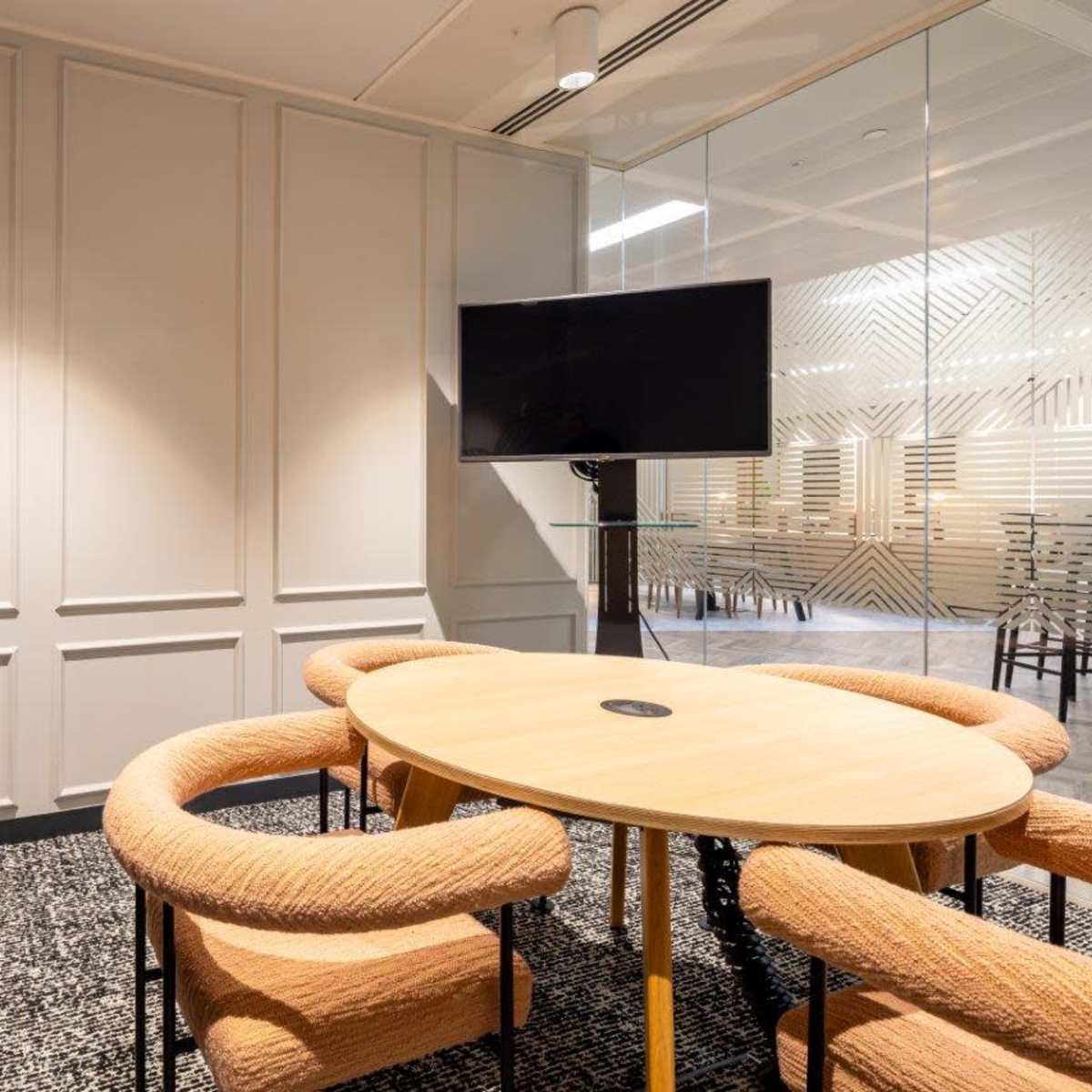 A modern conference room with a round wooden table surrounded by upholstered chairs, a glass wall in the background, and a television mounted on the wall.