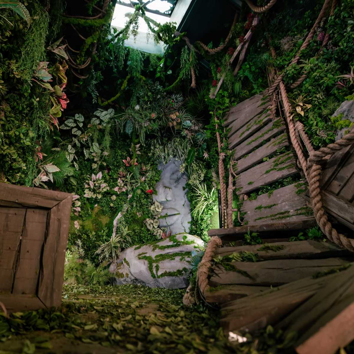A room filled with lush greenery, featuring a stone figure and wooden structures that appear to be in disarray.