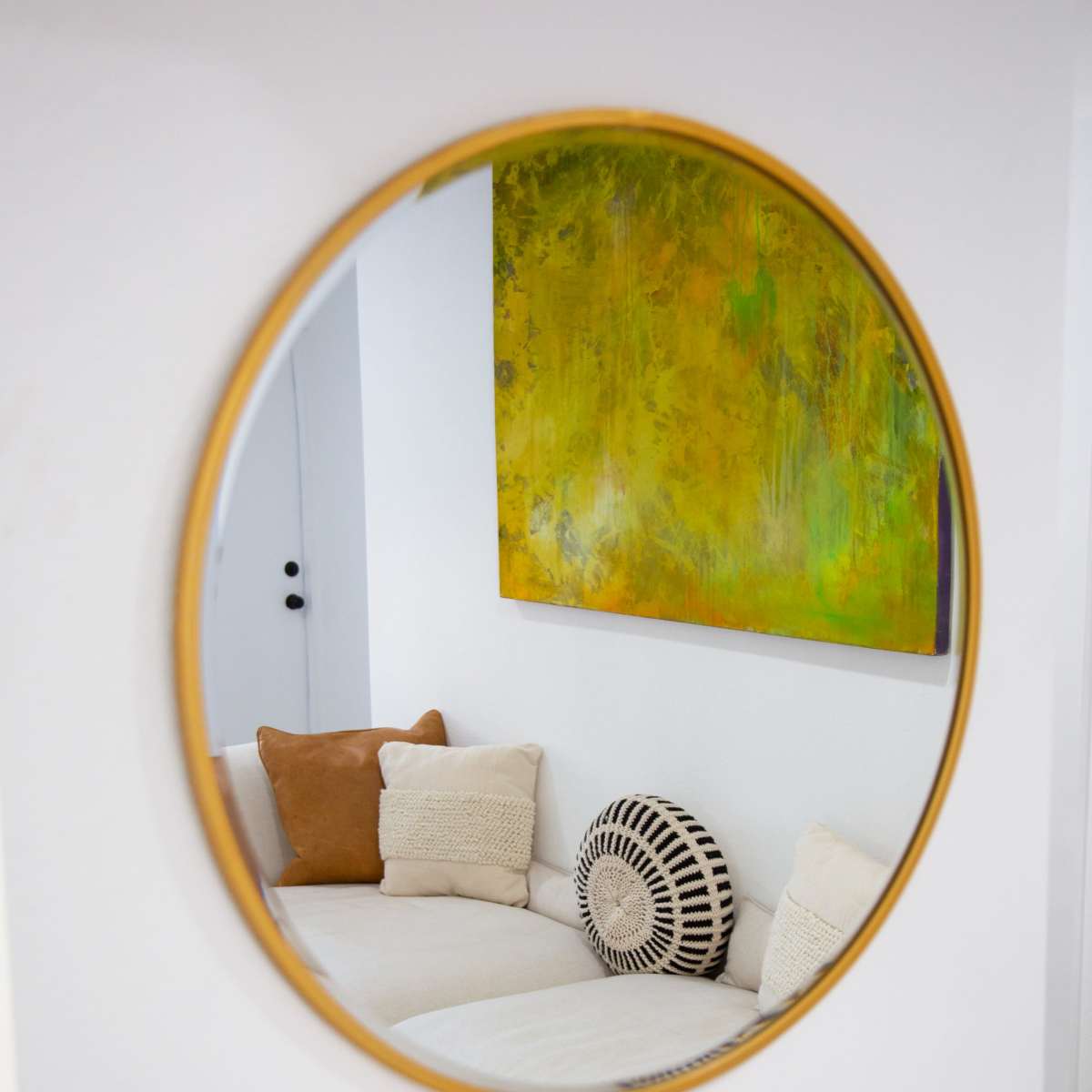 A round mirror reflects a beige couch with decorative pillows and a piece of green artwork on the wall.
