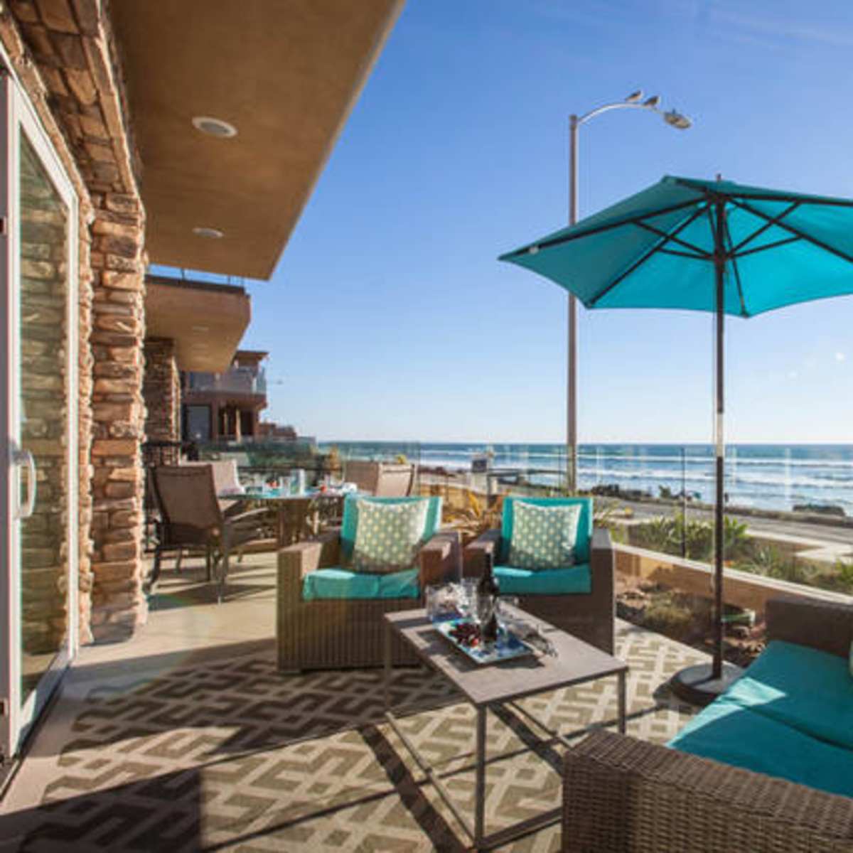 A patio area with turquoise umbrellas and chairs overlooks the ocean and waves under a clear blue sky.