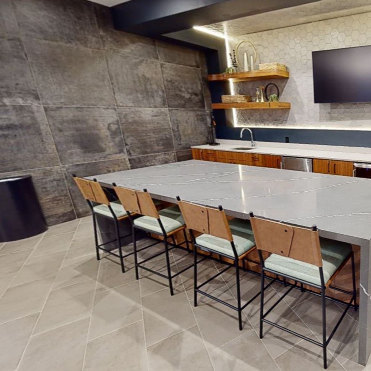 A modern kitchen with a large gray island, wooden cabinetry, a wall-mounted television, and hexagonal tile accents.