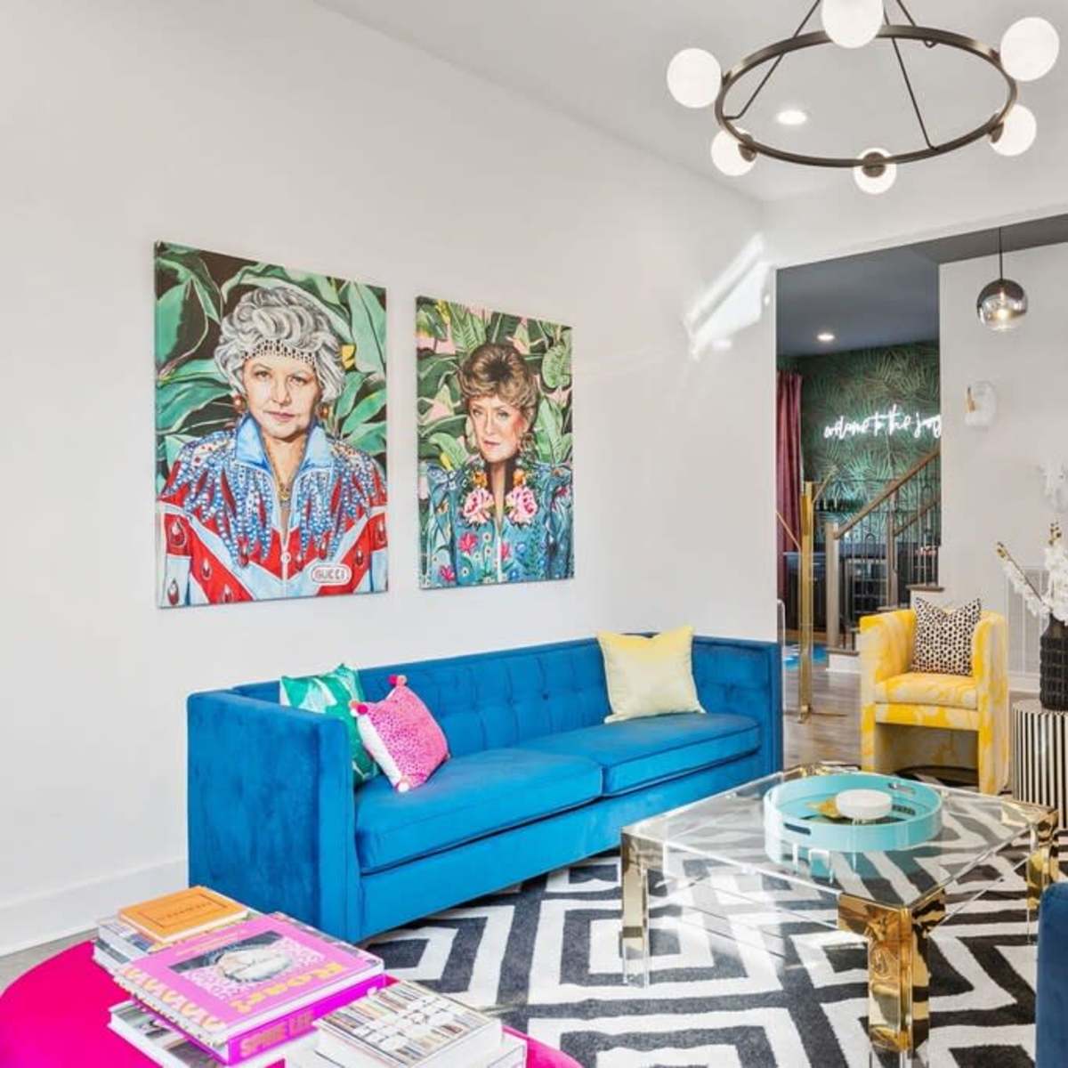 The living room features a mix of colorful furniture, including blue and yellow sofas, a coffee table, and large wall art depicting two women.