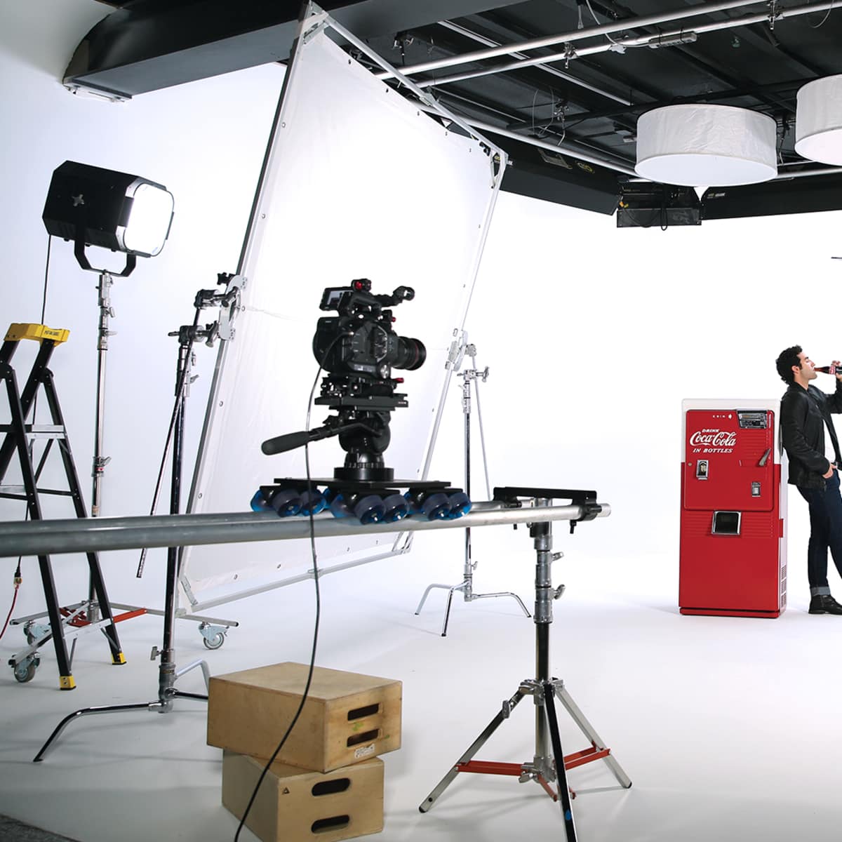 A man in a black suit stands by a red Coca-Cola vending machine in a photo studio filled with lighting equipment and backdrops.