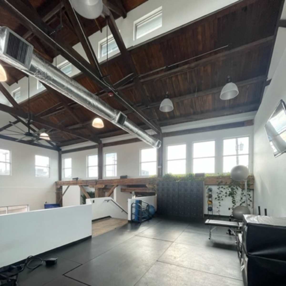 An open gym space with high wooden ceilings, large windows, and various fitness equipment arranged along the walls.