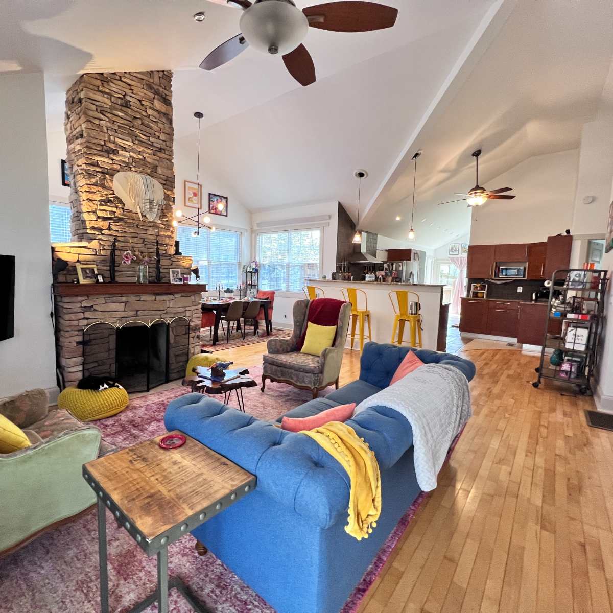 A living room with a stone fireplace, colorful furniture, and an open layout connecting to a dining area and kitchen.