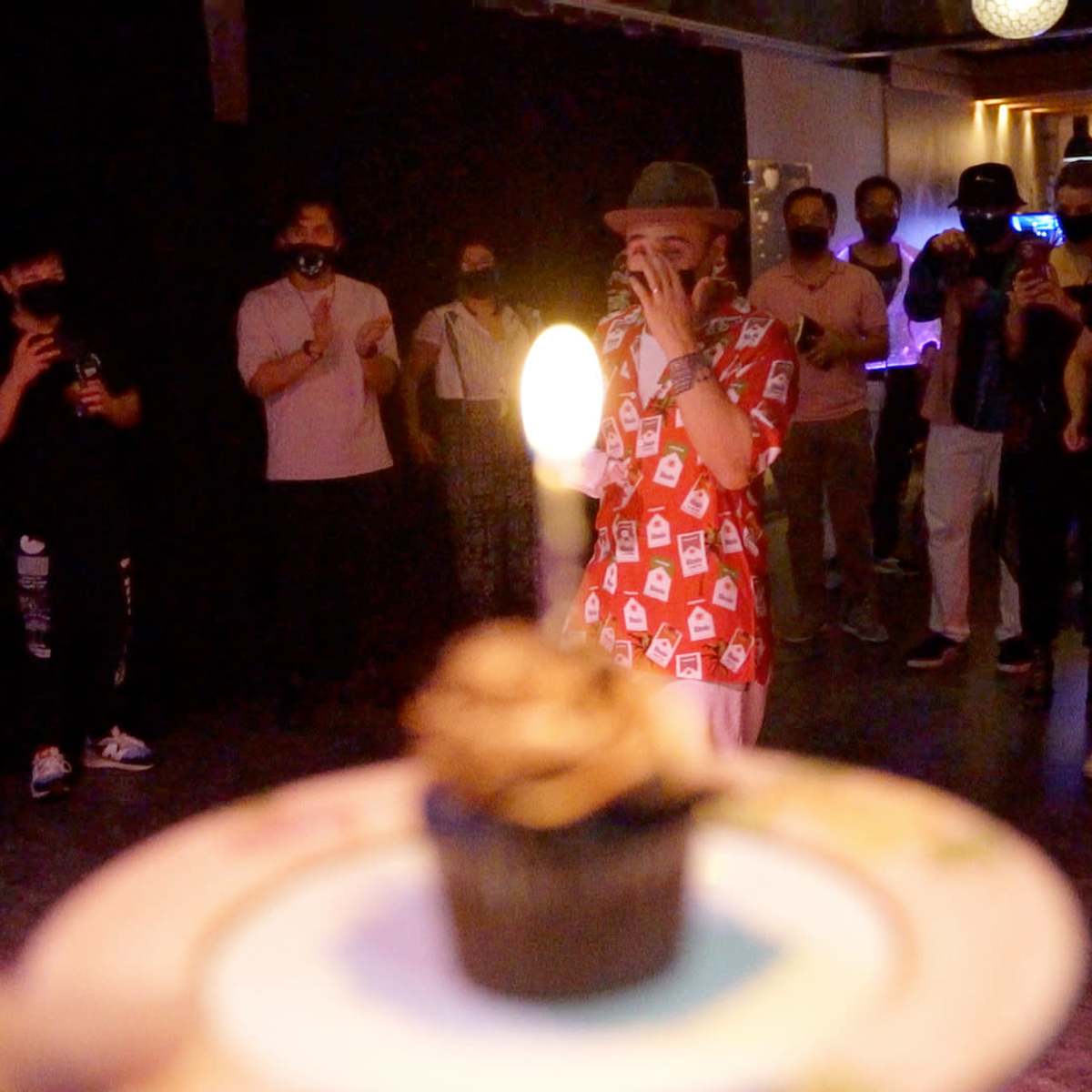 A person wearing a patterned shirt sings while holding a microphone in front of a group of people who are clapping and watching, with a cupcake in focus in the foreground.