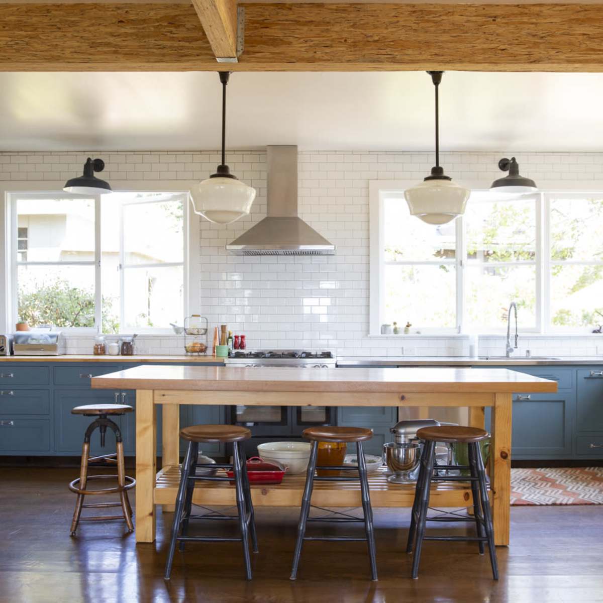 A modern kitchen features blue cabinetry, a central wooden island with stools, and large windows allowing natural light to brighten the space.