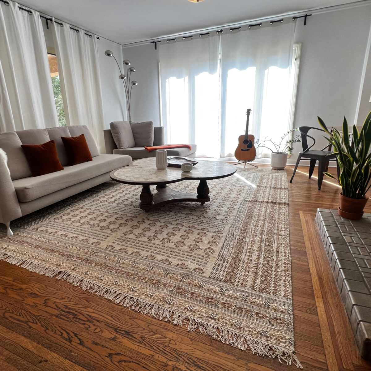 A light-filled living room with a sofa, a round coffee table, a guitar, and plants, all situated on a patterned area rug.
