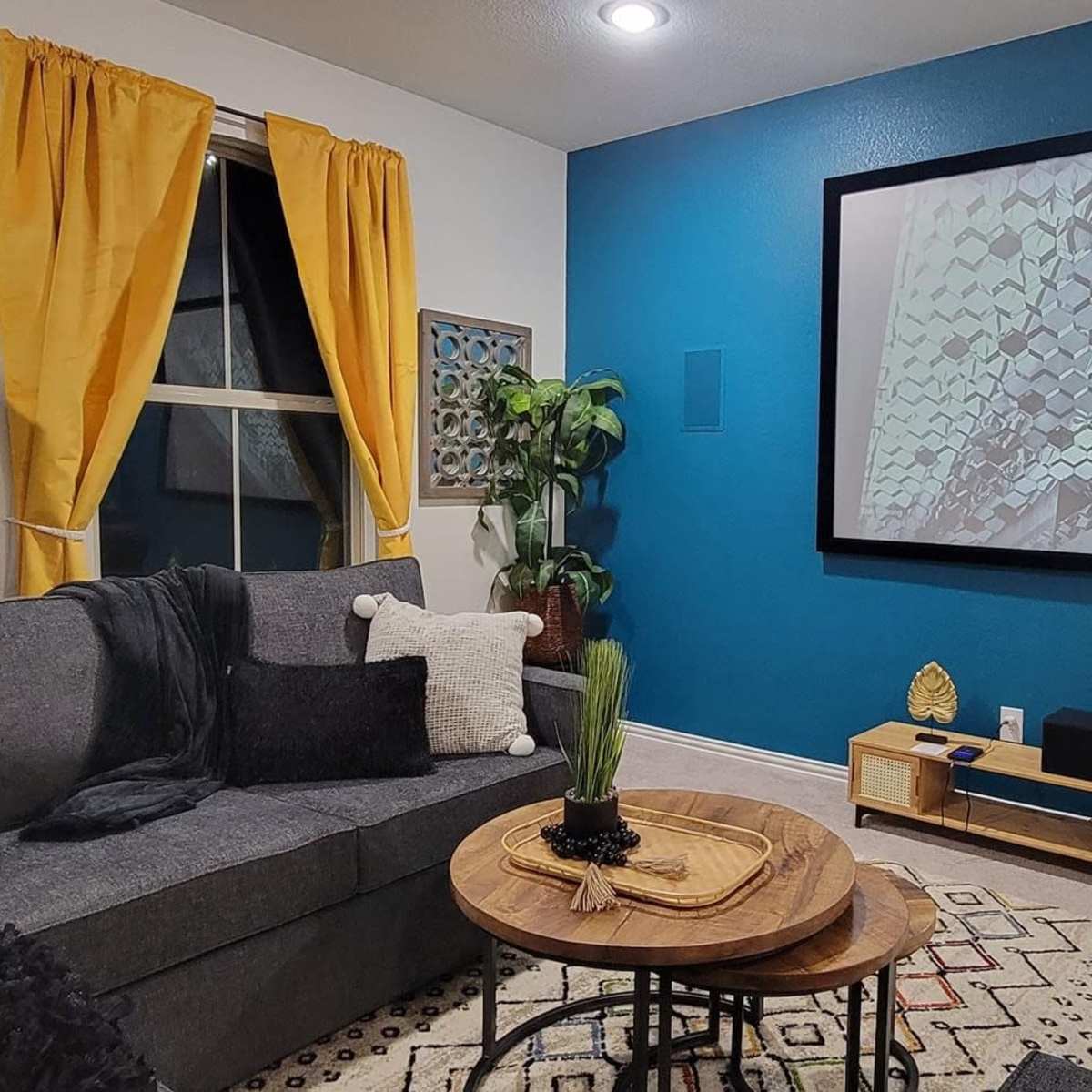 A living room features a large gray couch, a projector screen displaying a geometric pattern, and decorative plants against a blue wall with yellow curtains.