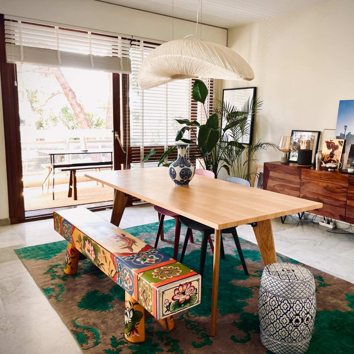 A dining room features a wooden table with a decorative vase, surrounded by colorful stools and a rug, against a backdrop of large windows and a plant.