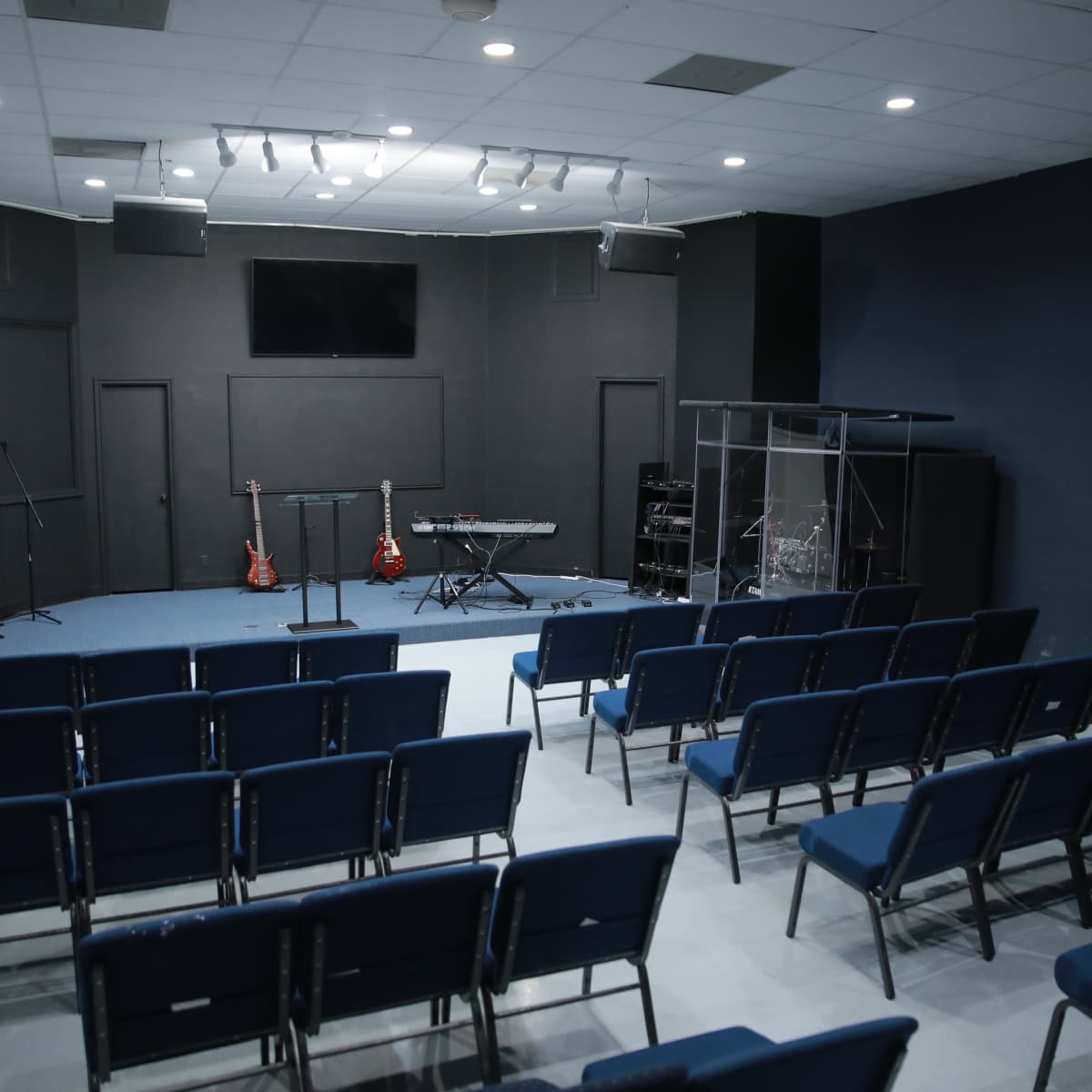 A music rehearsal room with rows of blue chairs facing a stage that features musical instruments and sound equipment.