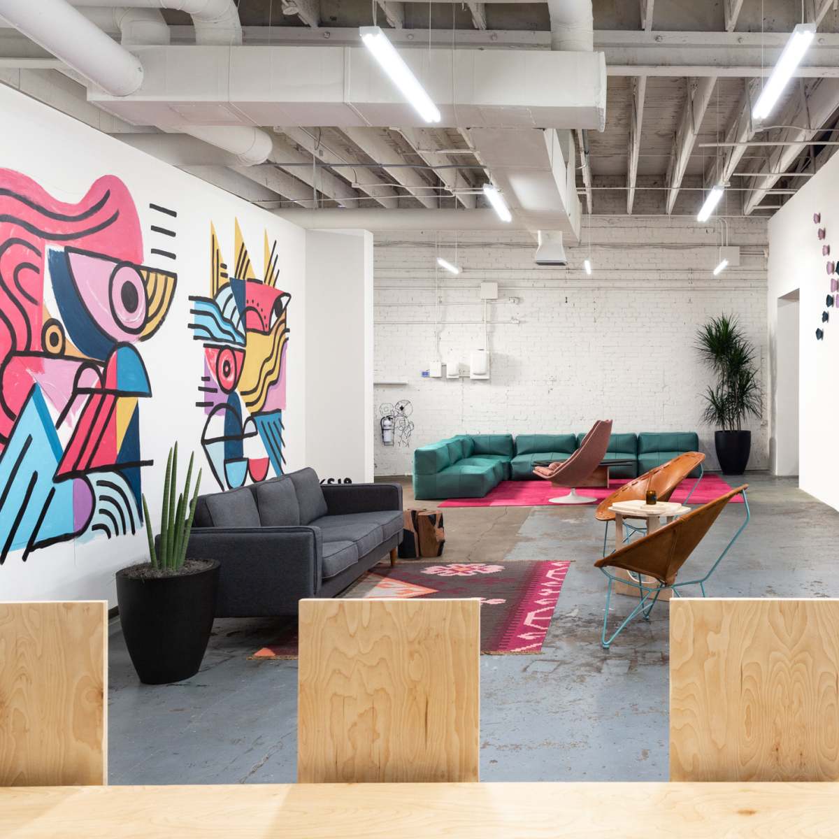A modern office space with colorful mural artwork, a lounge area featuring a gray sofa and green seating, and a large wooden table with black chairs.