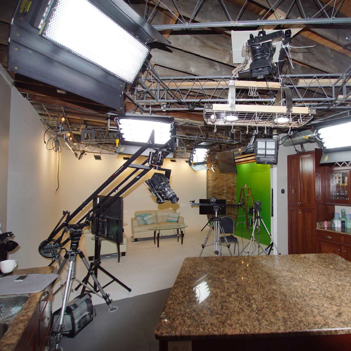 A television studio setup features multiple overhead lights, a green screen, and a kitchen island surrounded by cameras and equipment.