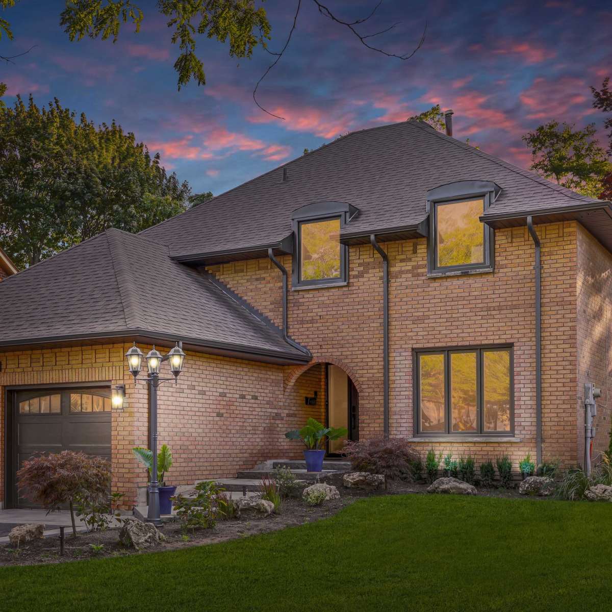A two-story brick house with a curved entrance and large windows is surrounded by landscaped gardens under a colorful sky at dusk.
