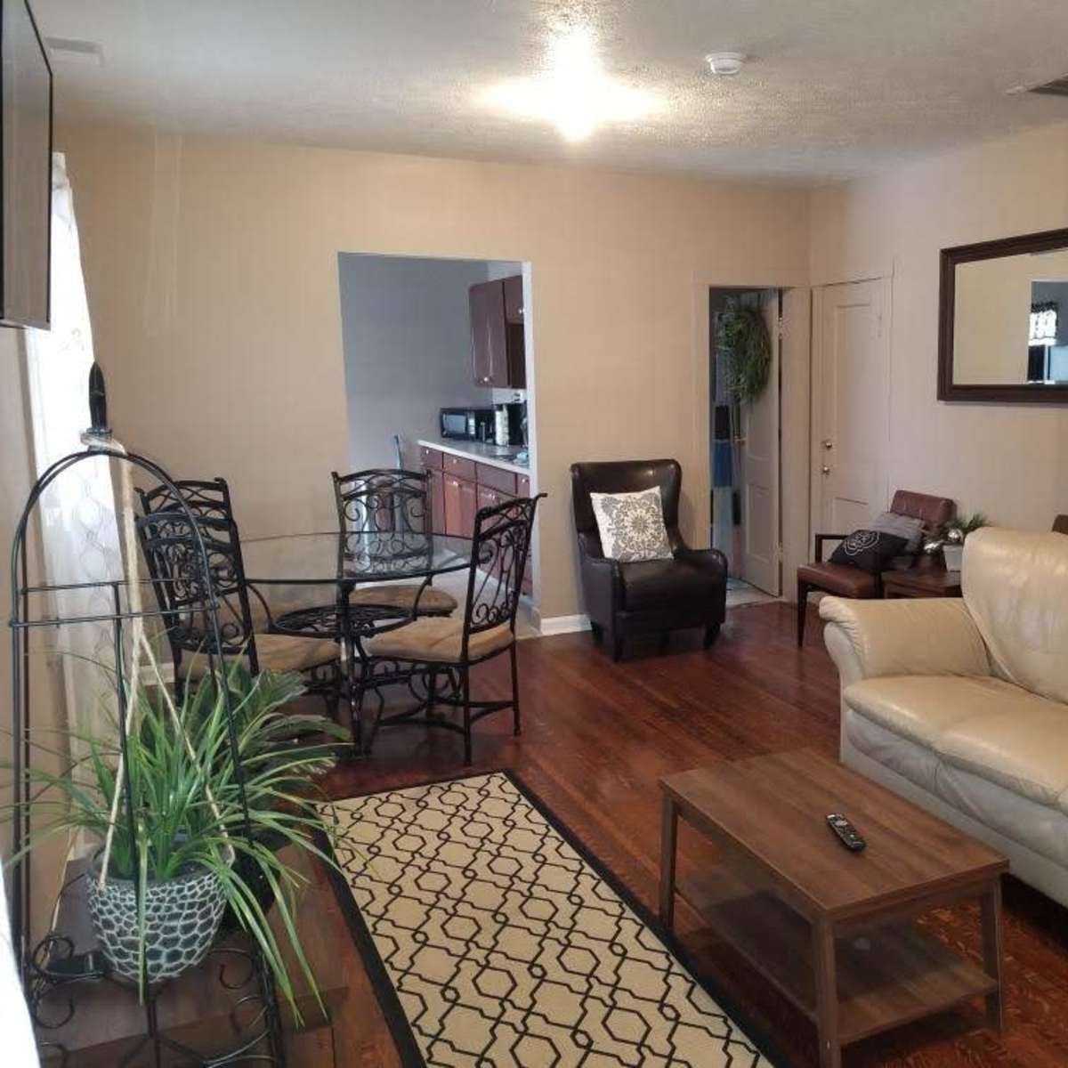 A living room with a leather sofa, a wooden coffee table, a dining area with a glass-top table and chairs, a large mirror on the wall, and a doorway leading to a kitchen.