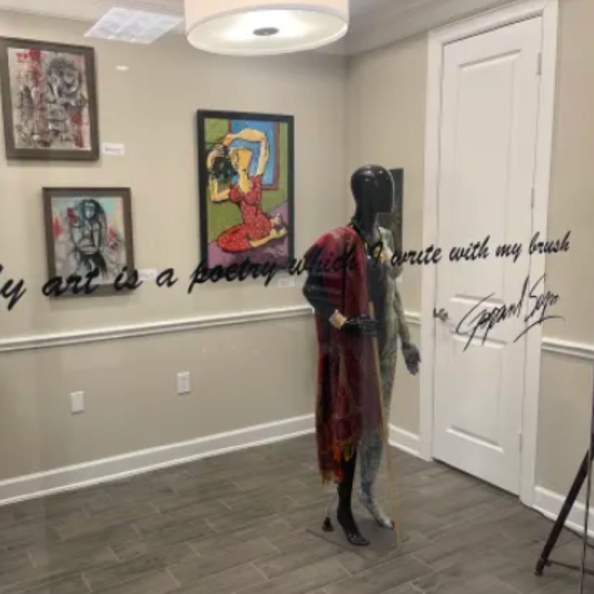A gallery space with framed artworks on the wall, a mannequin dressed in clothing, and a decorative quote displayed prominently.