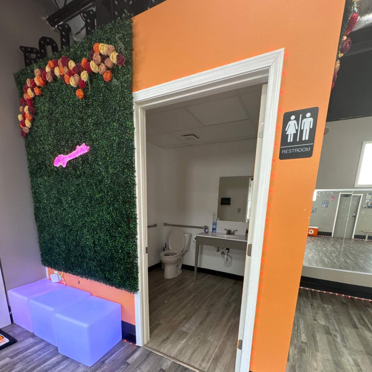 An entrance to a restroom, framed by a vibrant orange wall with decorative greenery and flowers, alongside a small waiting area with illuminated, translucent seats.
