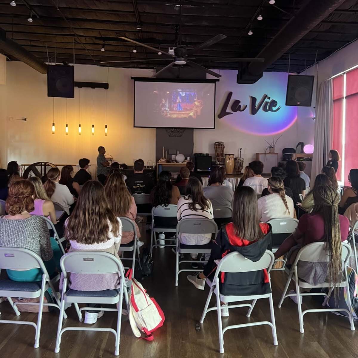 A group of people is seated in rows of chairs facing a large screen displaying a presentation in a well-lit room.