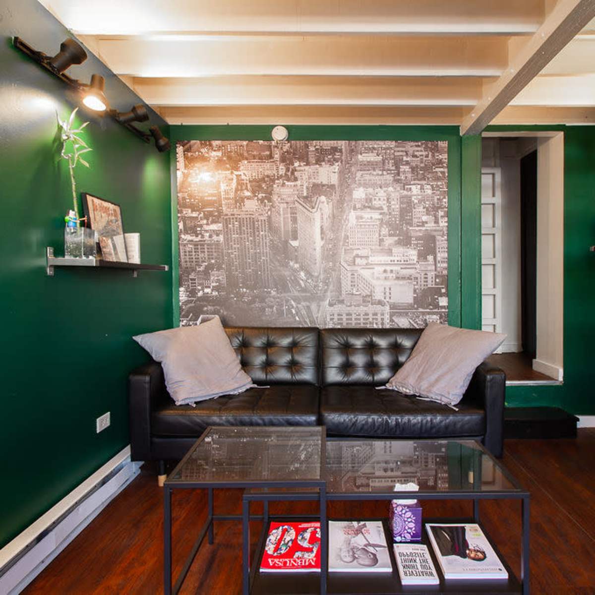 A small living space with dark green walls, a black leather couch, a coffee table with magazines, and shelves displaying various items.