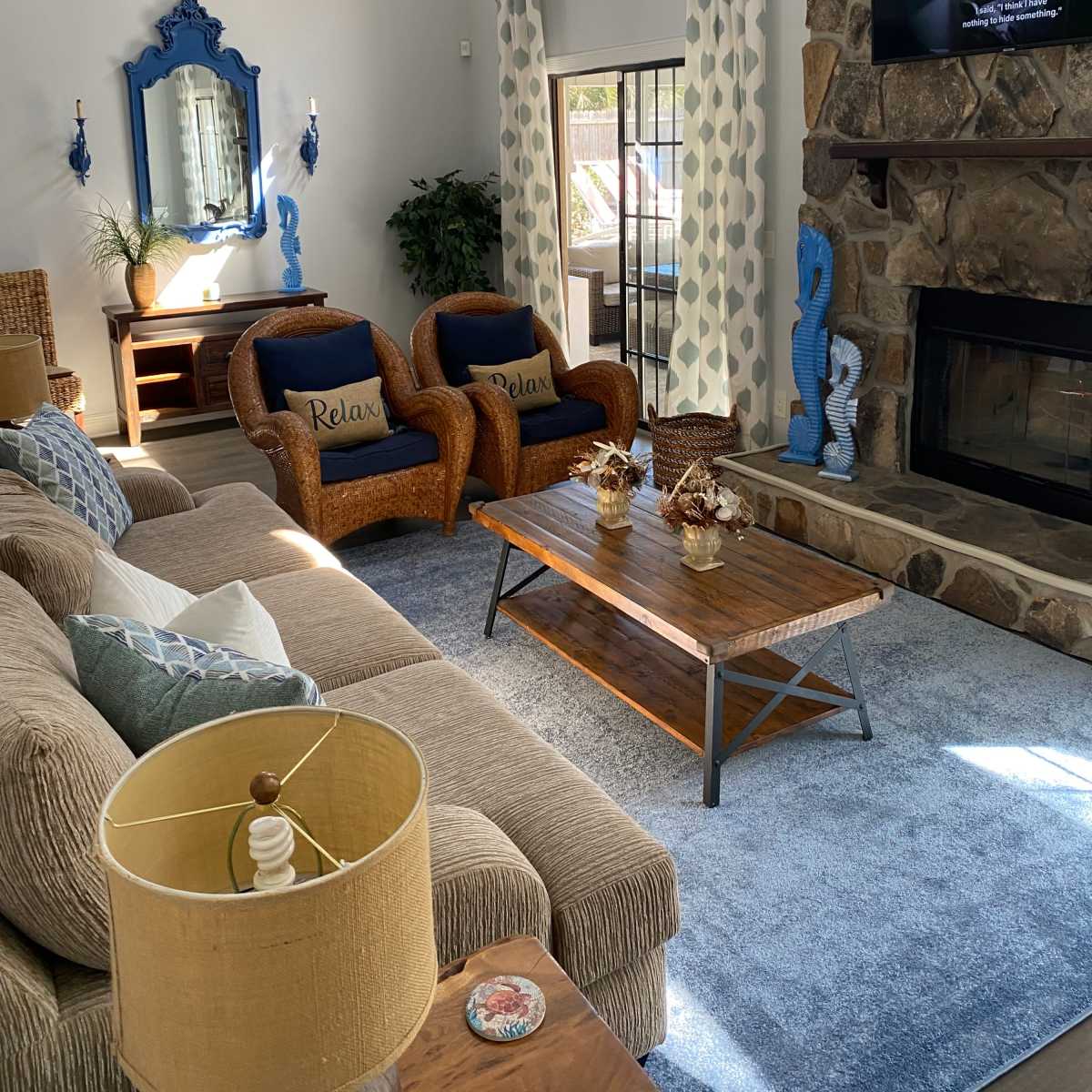 A cozy living room features a brown sectional sofa, two blue chairs with "Relax" cushions, a wooden coffee table, and a stone fireplace, with a large window allowing natural light to enter.