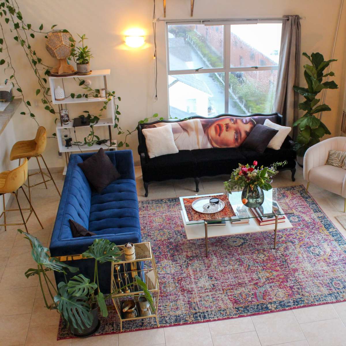 A modern living room features a blue sofa, a light-colored armchair, a coffee table with a floral arrangement, and various plants, all arranged around a colorful area rug.