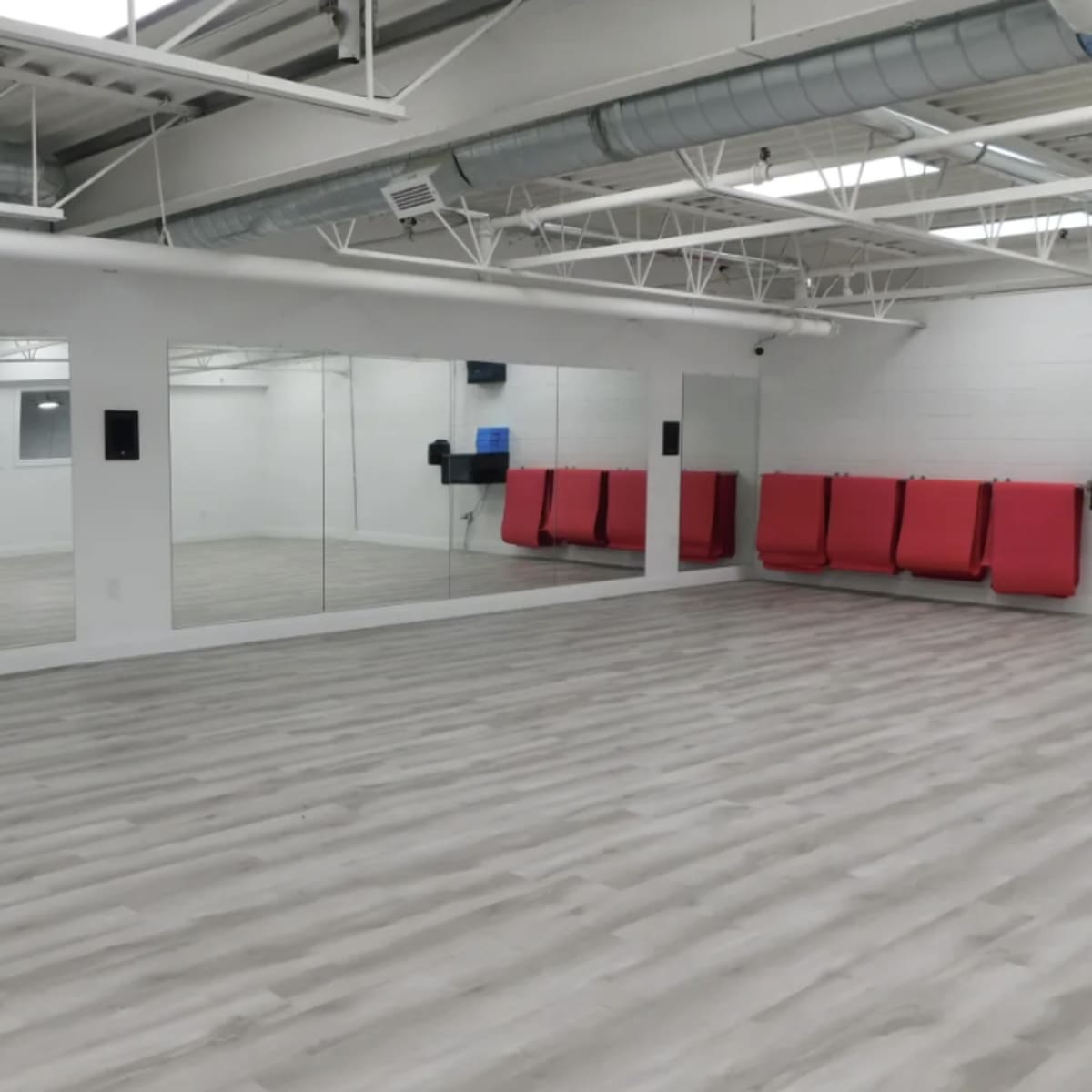A spacious, empty studio with light-colored wooden flooring, large mirrors on one wall, and red seating against another wall.