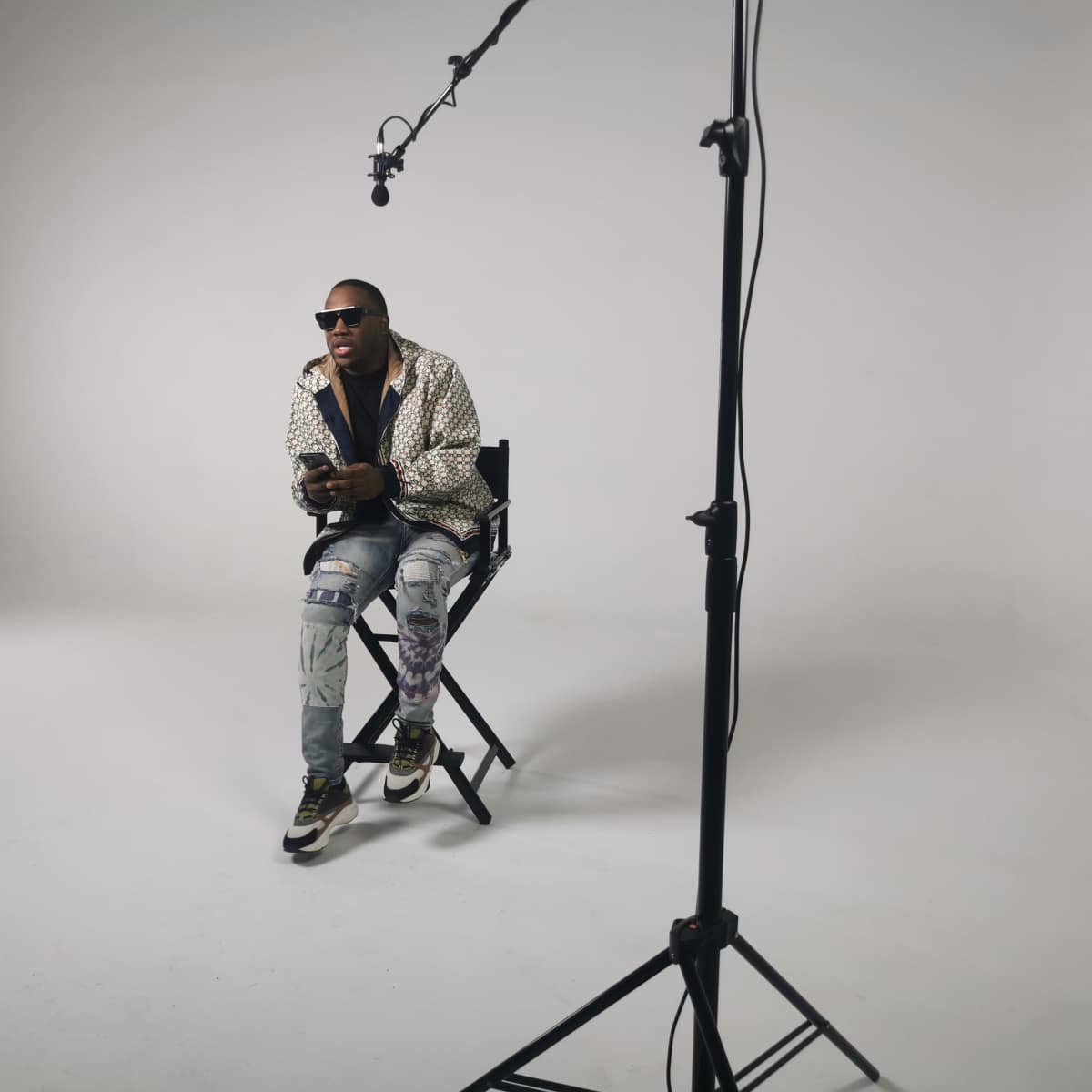 A person sits on a chair in a studio setting, with a microphone on a stand positioned above them and a plain backdrop.