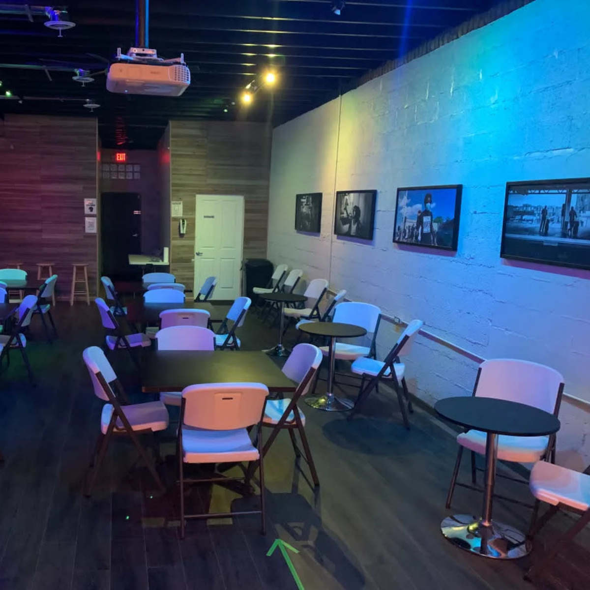The interior of a modern venue features several empty tables and chairs arranged in a spacious room with colorful lighting and photographs on the walls.