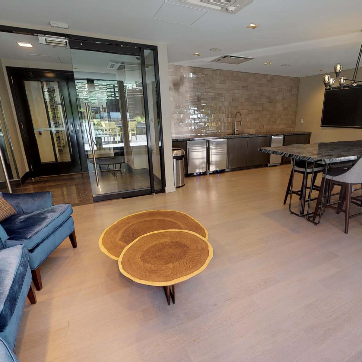 A modern common area with two blue chairs, a large table with seating, a kitchen space, and a glass door leading outside.