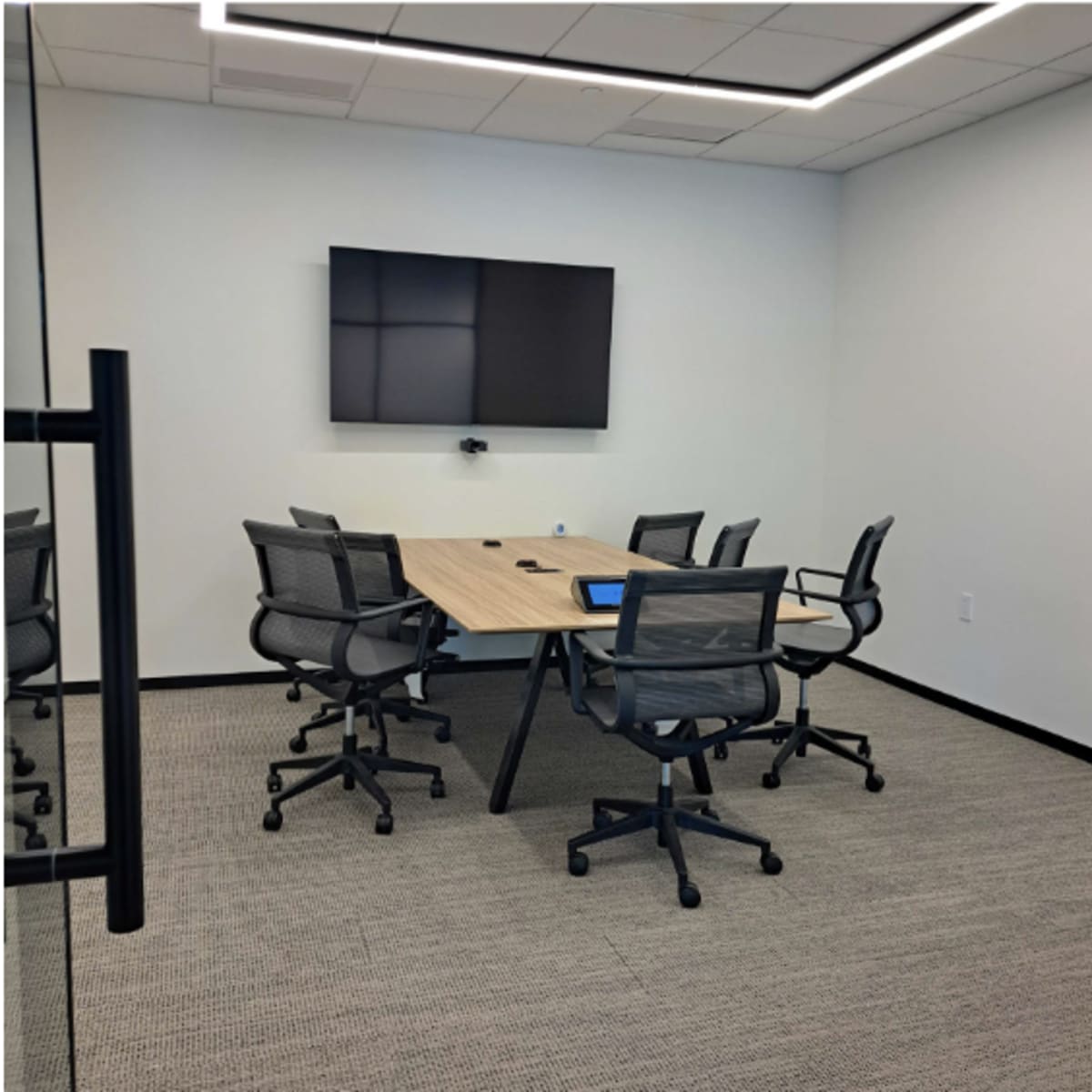 A modern, minimalist conference room with a rectangular table surrounded by six office chairs and a wall-mounted television.