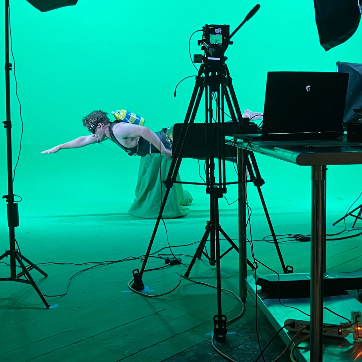 A person is swimming in an indoor studio with a green screen backdrop, surrounded by camera equipment and a laptop on a table.