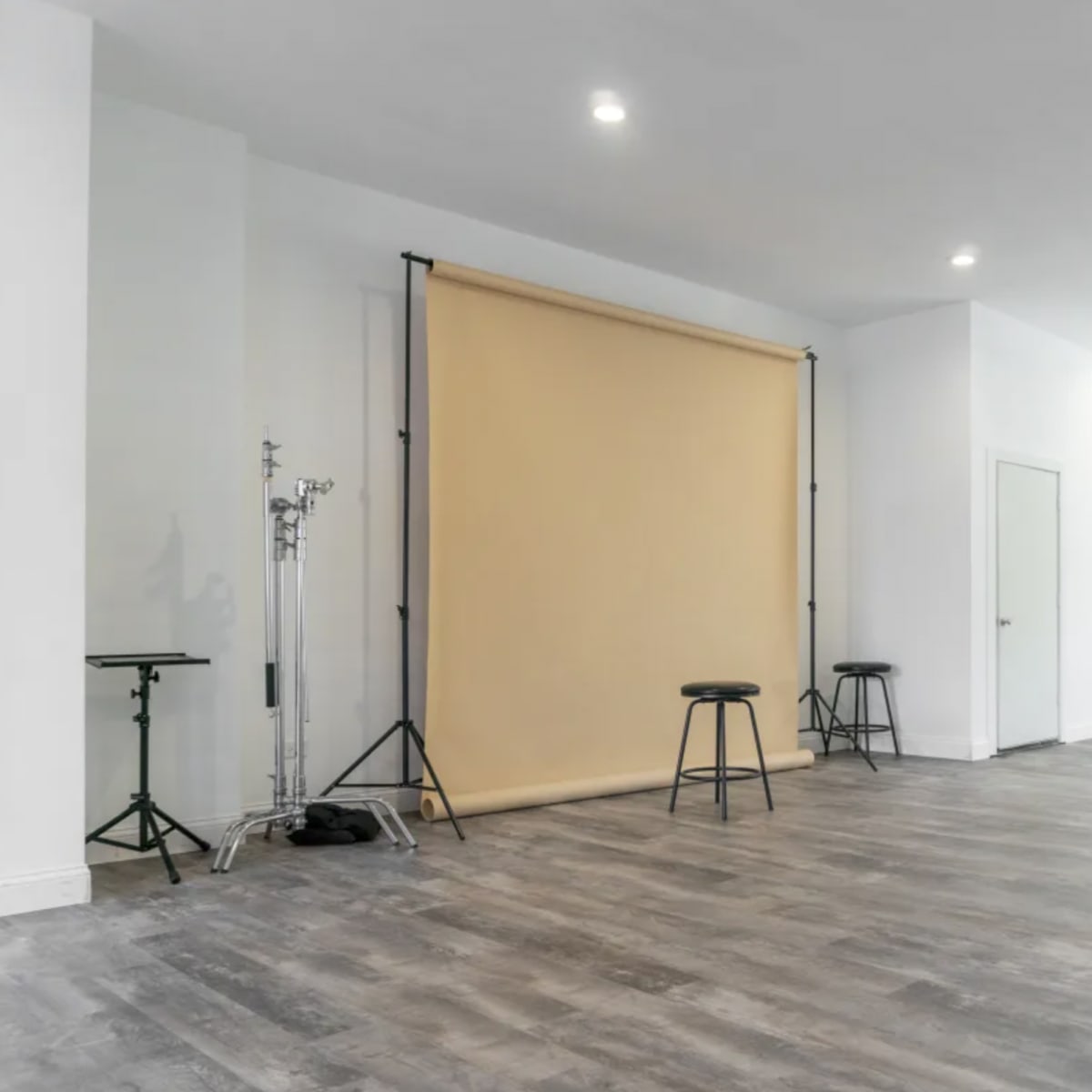 A spacious room with a beige backdrop set up on a stand, two black stools, and light-colored wooden flooring.