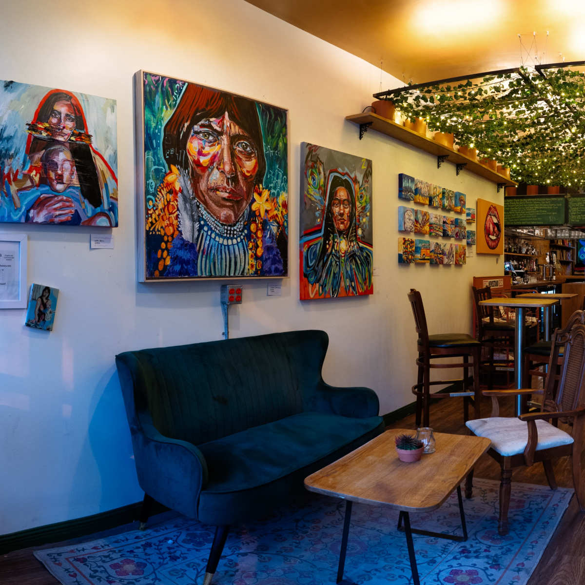 A cozy café interior featuring a green velvet sofa, wooden tables, and vibrant artwork on the walls, with overhead greenery creating a warm atmosphere.