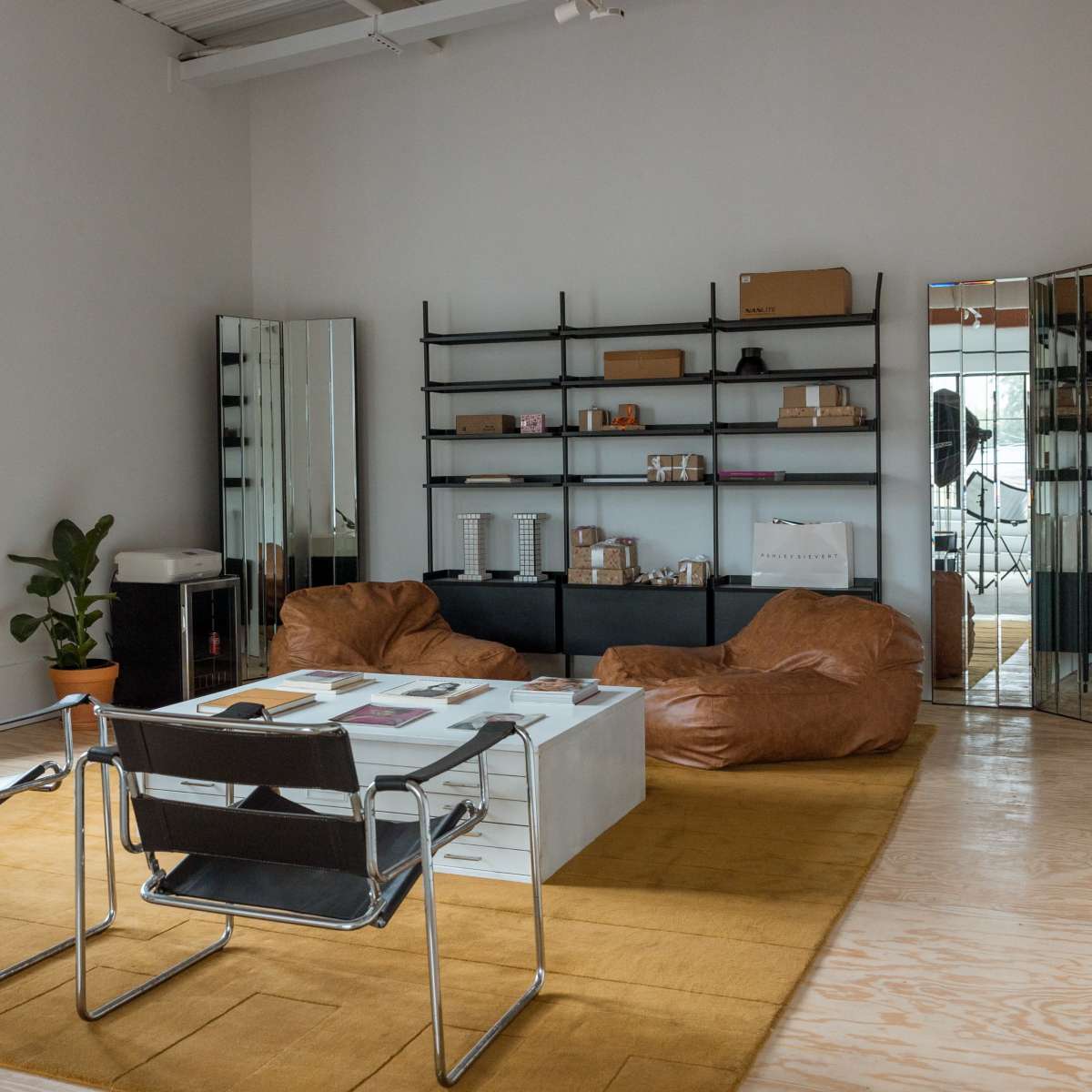 A modern room with a seating area featuring two chairs, a large table, and two bean bag chairs, alongside shelves filled with various items and a plant in the corner.