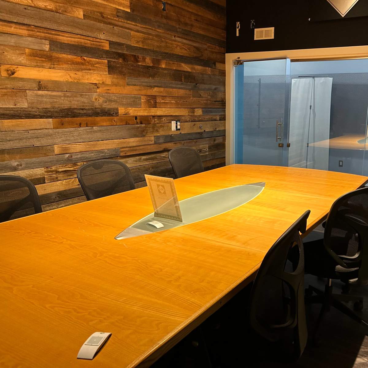 A modern conference room features a large wooden table with an oval glass centerpiece and black chairs arranged around it, adjacent to a glass-walled area.