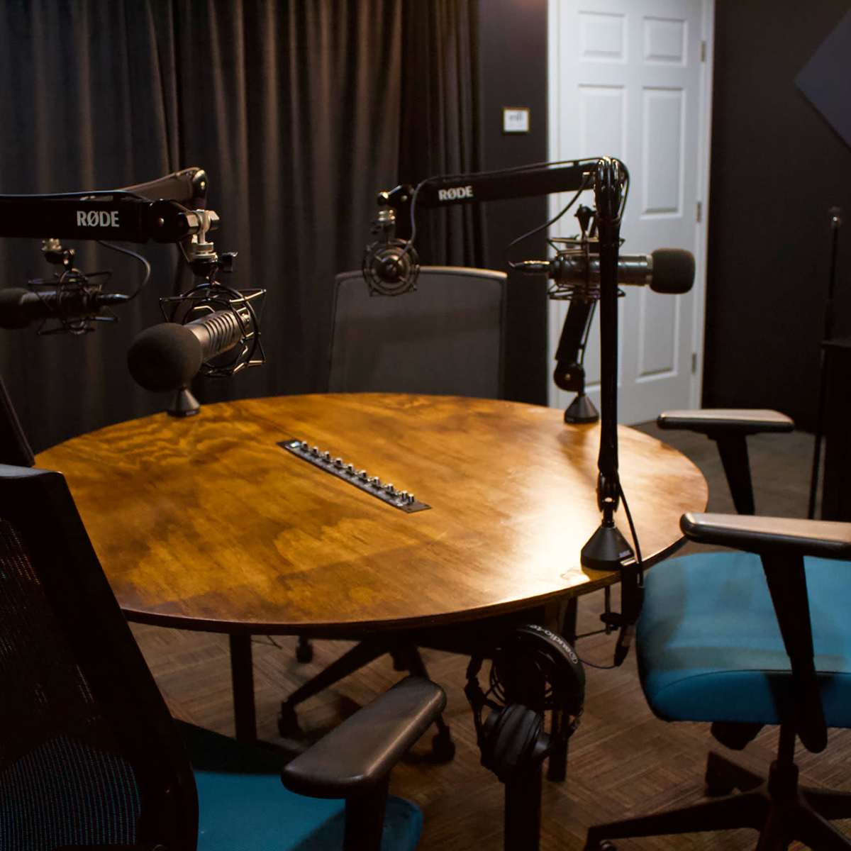 A round wooden table is surrounded by four chairs, with two microphone setups positioned above for recording.