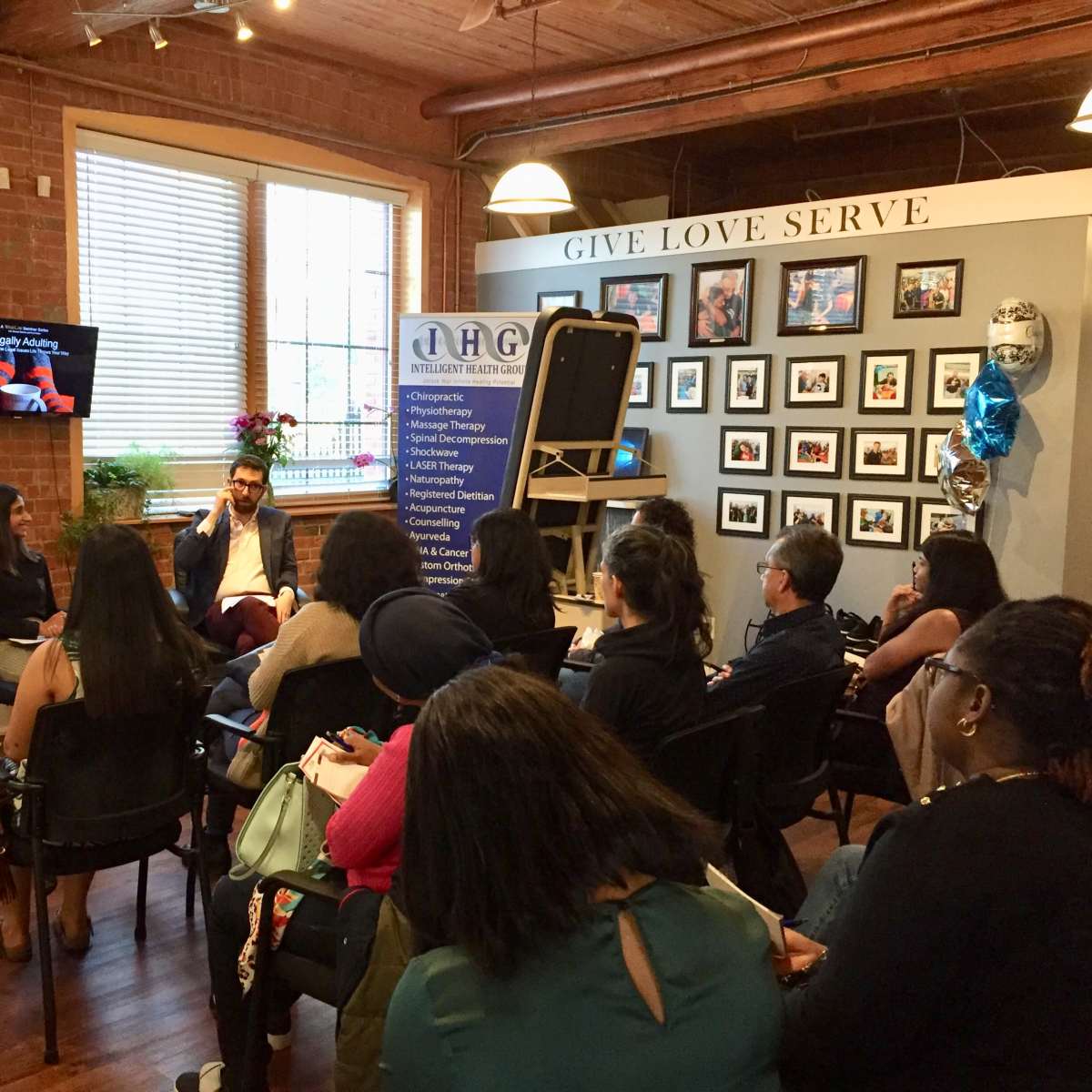 Two speakers engage with an audience in a room adorned with photographs and decor, while participants listen attentively from their seats.